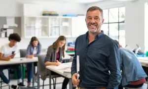 Un professore sorridente in aula, con studenti che studiano. Un esempio di come diventare professore superiori.
