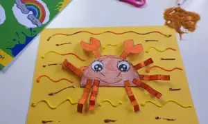 Granchio fatto a mano con carta e colla glitter, parte di lavoretti estivi. Forbici e colla glitter a lato.