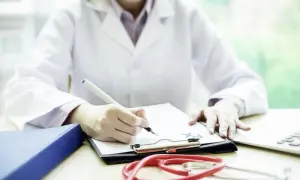 Medico legale che scrive su una cartella clinica, con stetoscopio e laptop. Quanto guadagna un medico legale?
