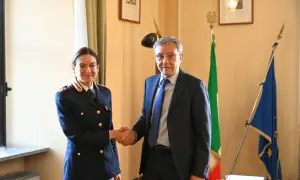 Il vice questore di Milano stringe la mano a un uomo in abito scuro, con le bandiere italiana ed europea sullo sfondo.