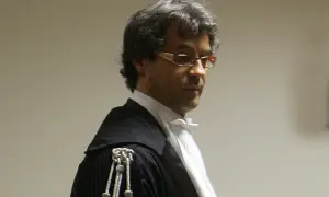 Un uomo con occhiali e toga nera, forse un sostituto procuratore o pm, guarda pensieroso.