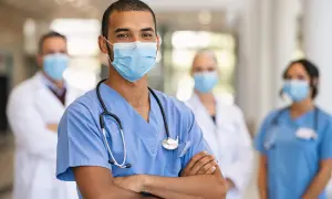 Un medico di pronto soccorso sorride con le braccia conserte, indossando una mascherina e uno stetoscopio. Dietro di lui, altri medici in camice bianco e blu. Quanto guadagna un medico di pronto soccorso?