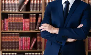 Uomo in abito blu con braccia conserte, circondato da scaffali pieni di libri legali. Un avvocato penalista pronto a difendere i suoi clienti.
