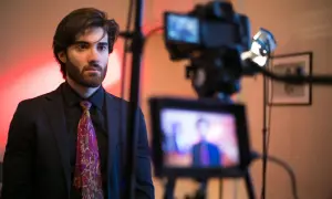Un giovane attore con barba e cravatta colorata, pronto per essere ripreso da una telecamera.