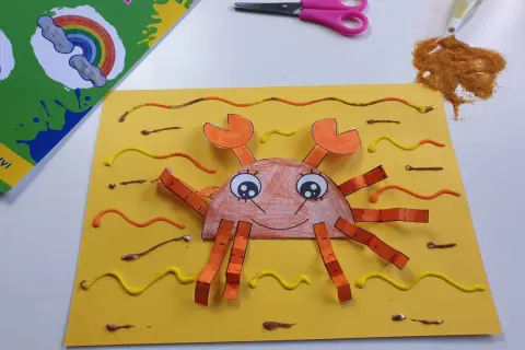 Granchio fatto a mano con carta e colla glitter, parte di lavoretti estivi. Forbici e colla glitter a lato.