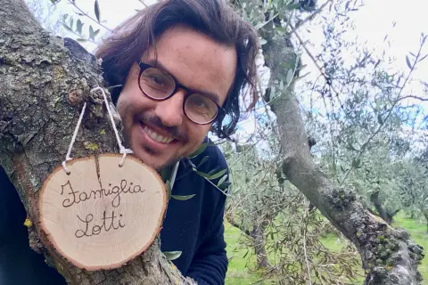 Il proprietario del b&b cerca personale. Sorride tra gli ulivi, con un cartello "Famiglia Lotti" appeso a un ramo.