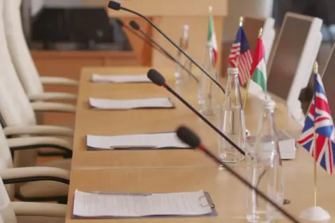Tavolo da conferenza con microfoni, bandiere di diverse nazioni e fogli. Forse qui si discute come si diventa console.