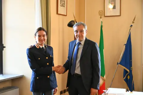 Il vice questore di Milano stringe la mano a un uomo in abito scuro, con le bandiere italiana ed europea sullo sfondo.