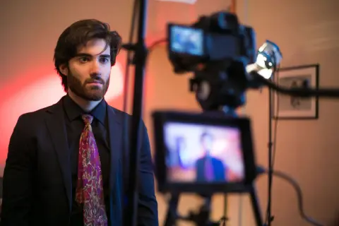 Un giovane attore con barba e cravatta colorata, pronto per essere ripreso da una telecamera.