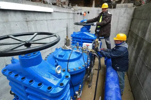 Lavoratori con elmetti gialli operano su grandi valvole blu di un acquedotto. Previste 200 assunzioni per il lavoro acquedotto pugliese.