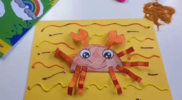 Granchio fatto a mano con carta e colla glitter, parte di lavoretti estivi. Forbici e colla glitter a lato.