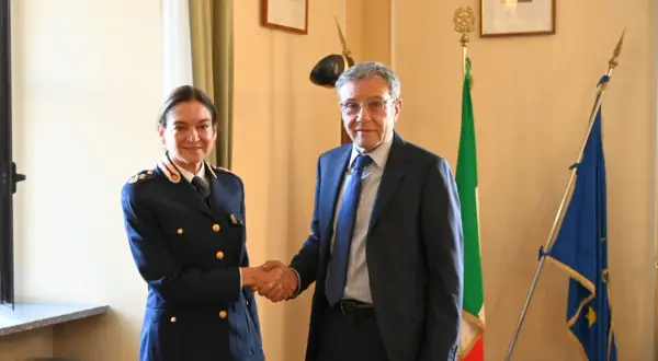 Il vice questore di Milano stringe la mano a un uomo in abito scuro, con le bandiere italiana ed europea sullo sfondo.