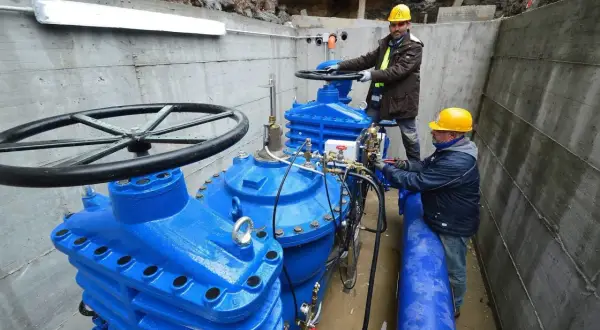 Lavoratori con elmetti gialli operano su grandi valvole blu di un acquedotto. Previste 200 assunzioni per il lavoro acquedotto pugliese.