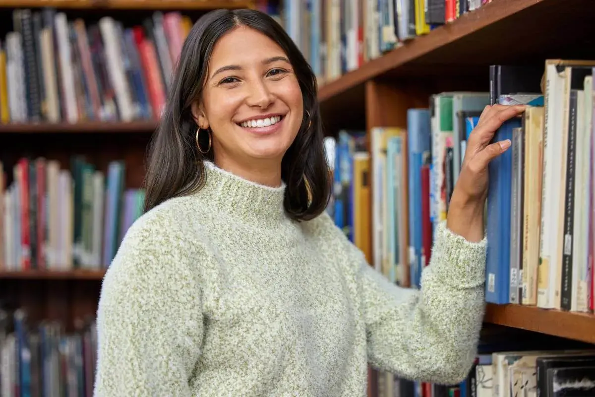 Sorriso sereno mentre sceglie un libro, immersa nell'atmosfera di chi ama lavorare in biblioteca.