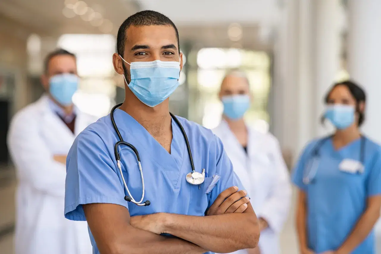Un medico di pronto soccorso sorride con le braccia conserte, indossando una mascherina e uno stetoscopio. Dietro di lui, altri medici in camice bianco e blu. Quanto guadagna un medico di pronto soccorso?