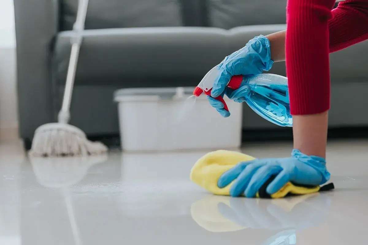Mani guantate spruzzano detergente e puliscono il pavimento. Inizia il lavoro pulizie mattina con un panno giallo e uno straccio sullo sfondo.
