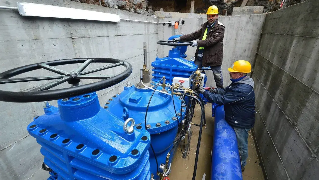 Lavoratori con elmetti gialli operano su grandi valvole blu di un acquedotto. Previste 200 assunzioni per il lavoro acquedotto pugliese.