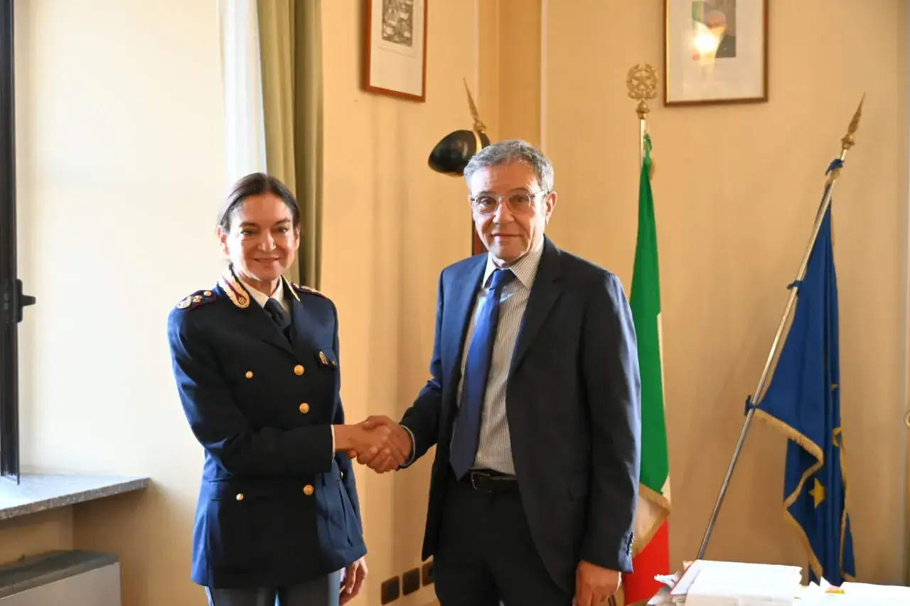 Il vice questore di Milano stringe la mano a un uomo in abito scuro, con le bandiere italiana ed europea sullo sfondo.