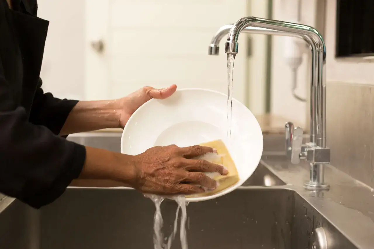 Mani che lavano un piatto sotto l'acqua corrente, con una spugna gialla. Un lavoro da lavapiatti part time, ma fatto con cura.