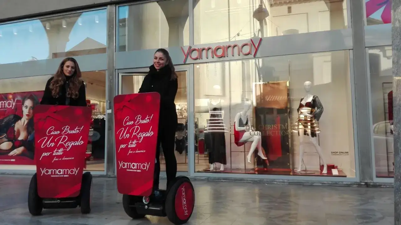 Promoter Yamamay su segway, con cartelloni pubblicitari, davanti al negozio. Promuove un regalo per tutti.