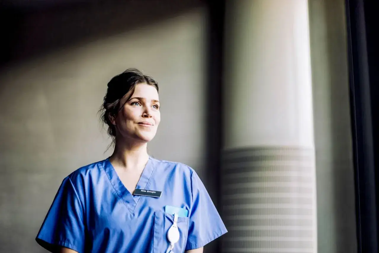 Donna con camice blu, sorride guardando fuori dalla finestra. Potrebbe essere un'infermiera in cerca di offerte lavoro OSS.