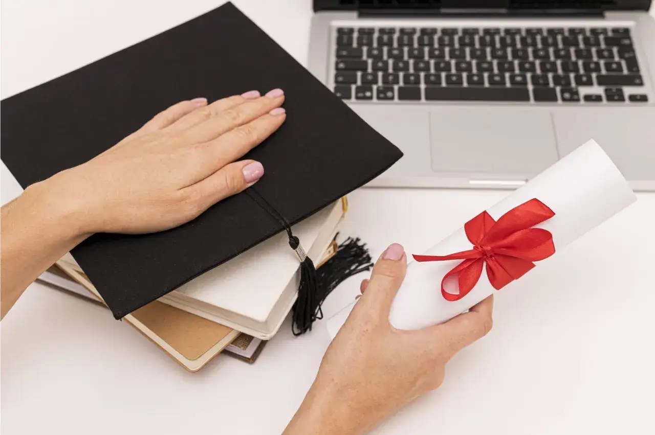 Mani che tengono un rotolo di diploma con fiocco rosso, accanto a un tocco di laurea e un laptop. Scopri come funziona la doppia laurea.