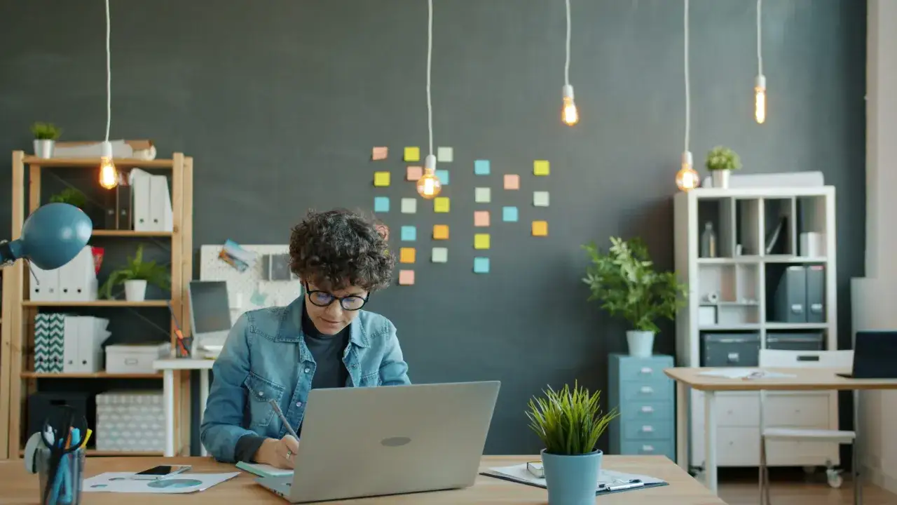 Ragazza con riccioli lavora da casa al suo laptop, circondata da post-it colorati e piante.