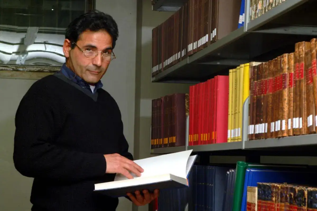 Uomo con occhiali legge un libro aperto, circondato da scaffali pieni di volumi antichi. Un momento di quiete mentre lavora in biblioteca.