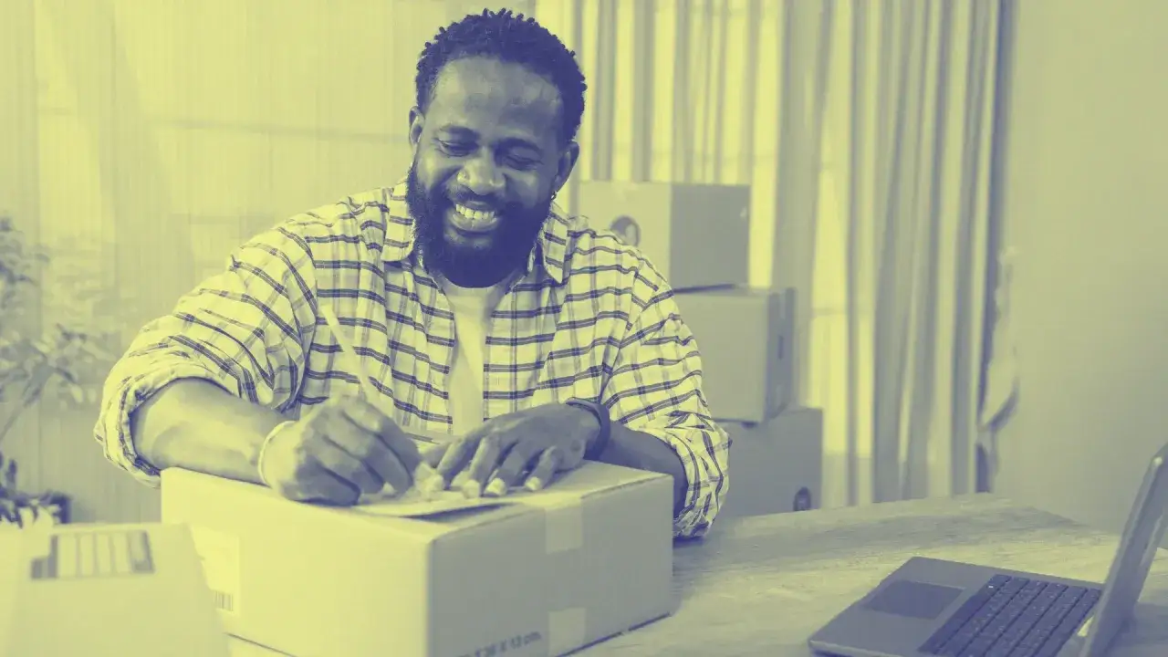 Uomo sorridente che prepara pacchi per il suo secondo lavoro da casa. Sul tavolo, un laptop e scatole in attesa di spedizione.