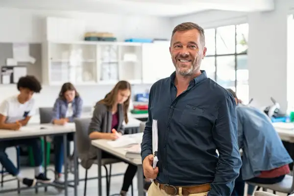 Un professore sorridente in aula, con studenti che studiano. Un esempio di come diventare professore superiori.