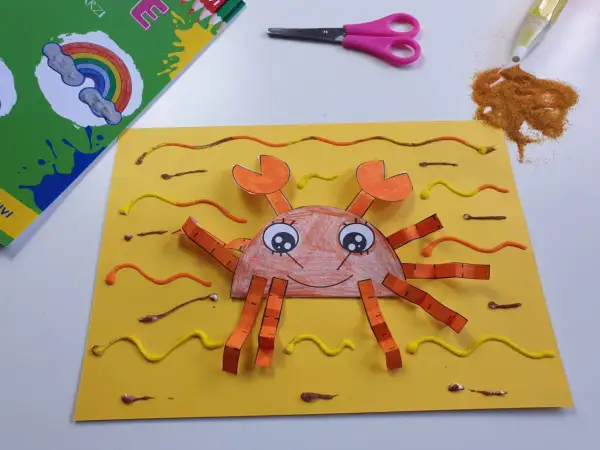 Granchio fatto a mano con carta e colla glitter, parte di lavoretti estivi. Forbici e colla glitter a lato.