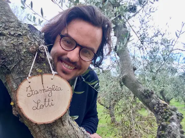 Il proprietario del b&b cerca personale. Sorride tra gli ulivi, con un cartello "Famiglia Lotti" appeso a un ramo.