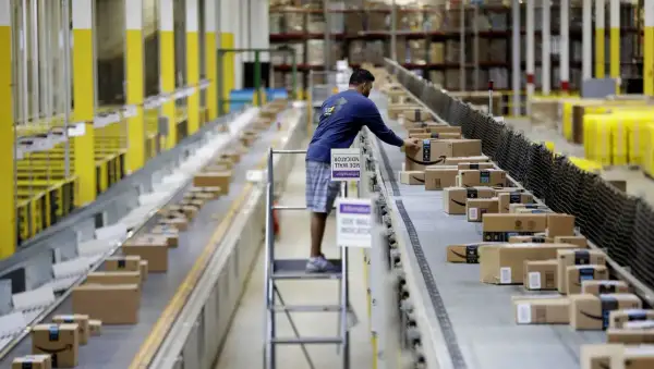 Un uomo lavora come corriere in un magazzino, sistemando pacchi su un nastro trasportatore pieno di scatole Amazon.