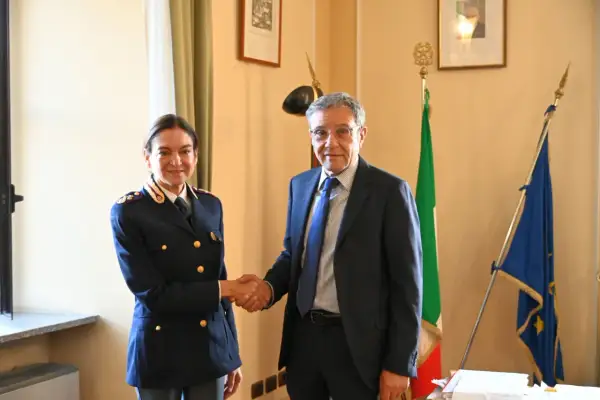 Il vice questore di Milano stringe la mano a un uomo in abito scuro, con le bandiere italiana ed europea sullo sfondo.