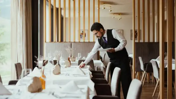 Cameriere in elegante divisa prepara la sala per la cena, un esempio di vitto e alloggio curato nei dettagli.