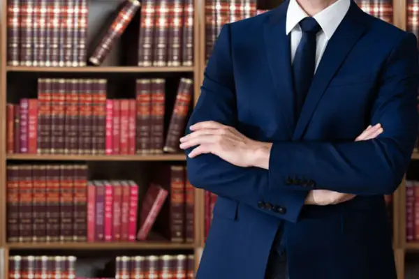 Uomo in abito blu con braccia conserte, circondato da scaffali pieni di libri legali. Un avvocato penalista pronto a difendere i suoi clienti.