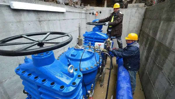 Lavoratori con elmetti gialli operano su grandi valvole blu di un acquedotto. Previste 200 assunzioni per il lavoro acquedotto pugliese.